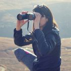 On a windswept hill a squatting woman peers through binoculars.