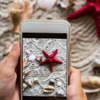 In front of a red sea star and shells on a beach hands hold a smart phone that has a picture of the scene on its screen