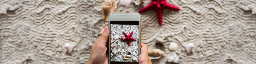 In front of a red sea star and shells on a beach hands hold a smart phone that has a picture of the scene on its screen