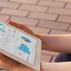 Against the background of a brick patio, a seated woman holds a white tablet displaying a Power BI dashboard